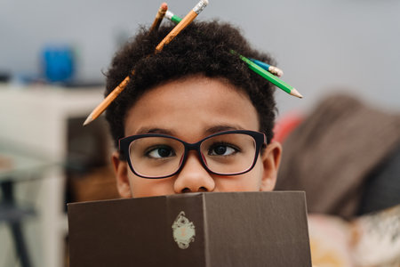 Black boy making fun with pencils while reading book at homeの写真素材