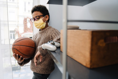 Black boy in face mask holding basketball while standing at homeの写真素材
