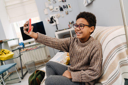 Black boy in eyeglasses taking selfie on cellphone while resting at homeの写真素材