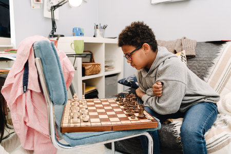Black boy in eyeglasses playing chess while sitting on sofa at homeの写真素材