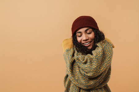 Young black woman wearing hat laughing and hugging herself isolated over beige backgroundの写真素材