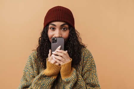 Surprised black woman wearing hat using mobile phone isolated over beige background