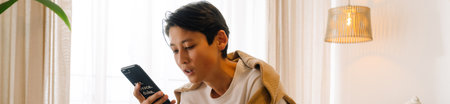 Asian boy using mobile phone while sitting on bed at homeの写真素材