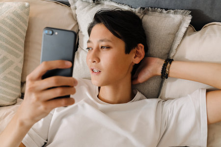 Asian boy using mobile phone while resting on bed at homeの写真素材