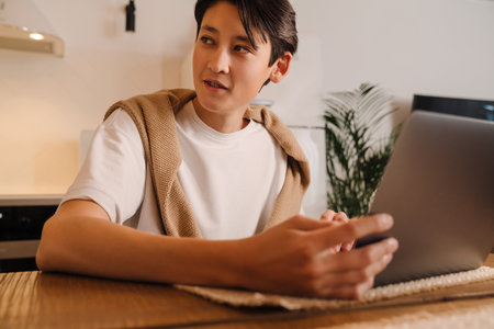 Asian boy working with laptop while sitting in kitchen at homeの写真素材