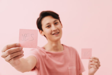 Asian boy wearing t-shirt smiling while showing sticker at camera isolated over pink backgroundの写真素材