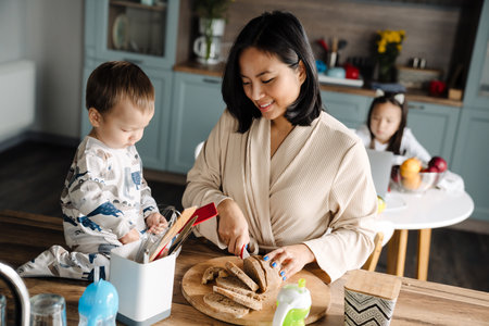 Happy mother smiling and cutting bread while spending time with her children at homeの写真素材