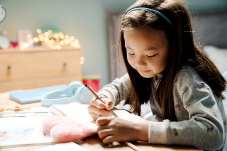 Asian girl writing in exercise book while doing homework at table at homeの写真素材