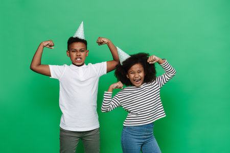 Black boy and girl grimacing while showing their biceps together isolated over green background