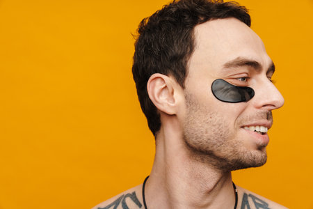 Close up of a shirtless mid aged brunette man wearing cosmetic eye patches over yellow wall...