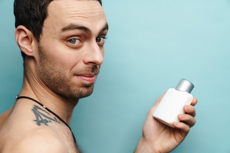 Shirtless white man smiling while holding cologne isolated over blue wallの写真素材