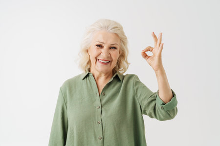Grey senior woman in shirt smiling and gesturing ok sing isolated over white background