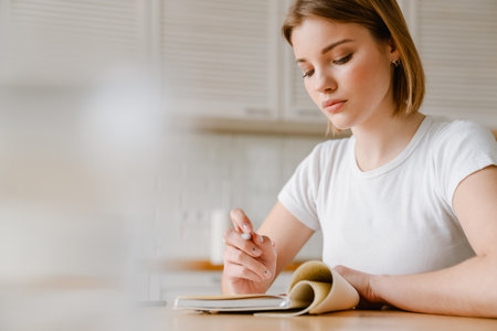 Smiling young woman writing in her diary while sitting at the kitchen table at homeの写真素材
