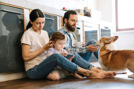 Smiling young family sitting on a floor of a kitchen at home using mobile phone playing with dogの写真素材