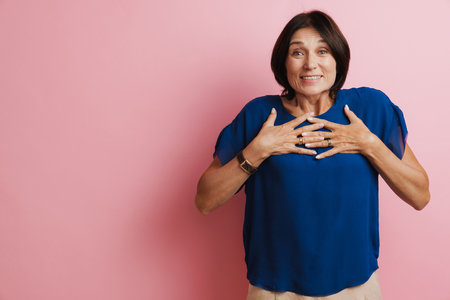 Mature brunette woman smiling while holding hands on her chest isolated over pink backgroundの写真素材