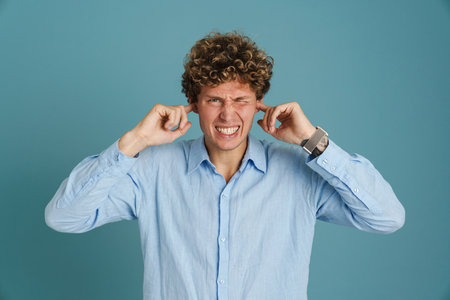 Young curly man wearing shirt frowning and plugging her ears isolated over blue background