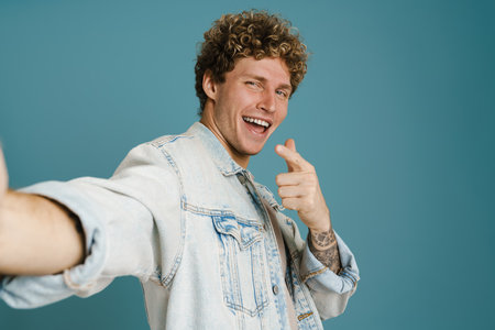 Young excited man pointing finger away while taking selfie photo isolated over blue background