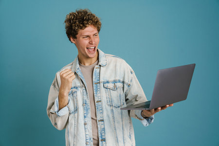 Young man making winner gesture while posing with laptop isolated over blue background