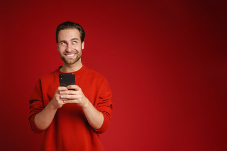 Young bristle man wearing sweater smiling and using cellphone isolated over red backgroundの写真素材