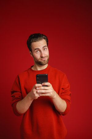 Young perplexed man looking at camera and using cellphone isolated over red backgroundの写真素材