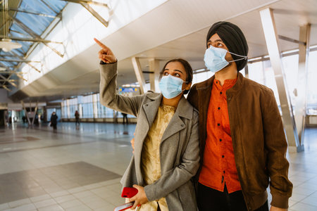 Indian couple wearing face masks hugging and pointing finger away in airport indoorsの写真素材