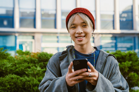 Asian boy smiling while using mobile phone and earphones on city street