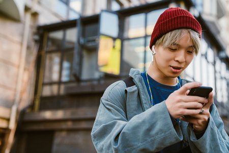 Asian boy wearing hat using mobile phone and earphones on city streetの写真素材