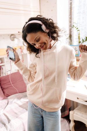 African happy teen in headphones listening to music and dancing in her cozy room at homeの写真素材