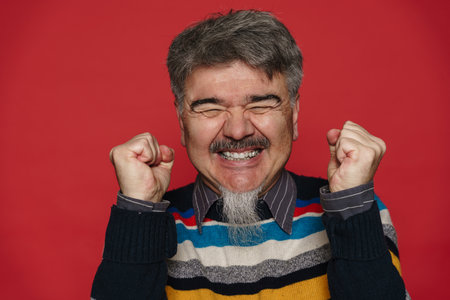 Mature man with grey beard smiling and making winner gesture isolated over red background