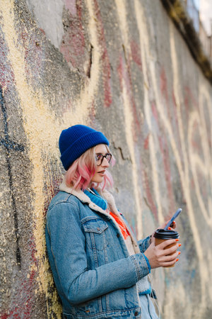 Young woman using cellphone and drinking coffee while standing by wall outdoorsの写真素材