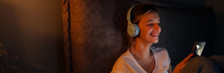 Young woman in headphones smiling and using cellphone while sitting on bed at homeの写真素材