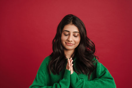 East asian woman smiling and holding palms together isolated over red background