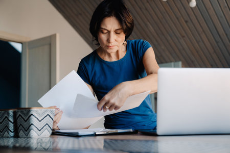 White mature woman working with laptop and papers in kitchen at homeの写真素材
