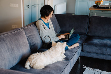 White mature woman using laptop while sitting with her dog on couch at homeの写真素材