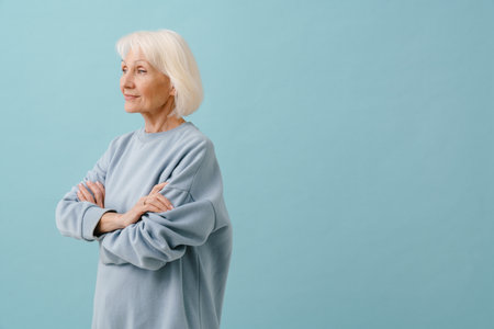 Beautiful senior woman in blue pullover standing with her left side with folded arms over isolated blue backgroundの写真素材
