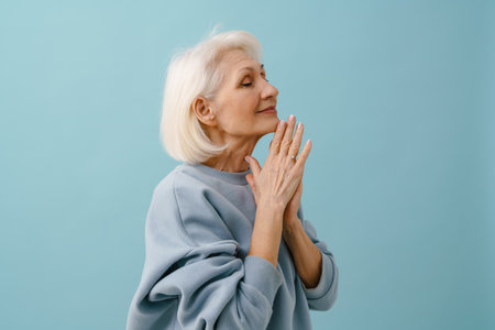 Beautiful senior woman in blue pullover with closed eyes and folded hands over isolated blue...
