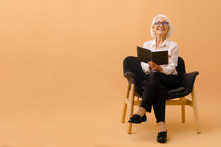 Beautiful smiling senior woman in business suit sitting on armchair with notebook over isolated brown backgroundの写真素材