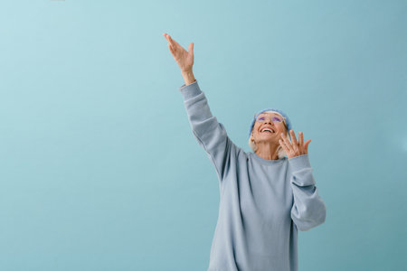 Beautiful enthusiastic senior woman in winter hat and glasses with raised arms looking upward over isolated blue backgroundの写真素材