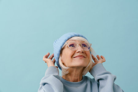 Beautiful smiling senior woman in glasses adjusting her winter hat and looking upward over isolated blue backgroundの写真素材