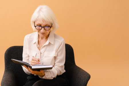Beautiful smiling senior woman in business suit sitting on armchair and writing notes over isolated brown backgroundの写真素材
