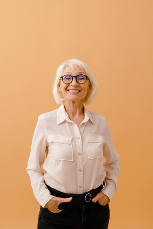 Beautiful happy smiling senior woman in business suit and glasses standing over isolated brown...