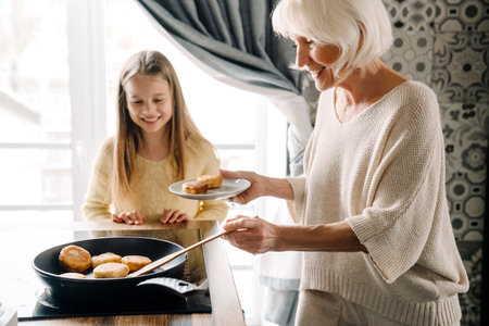 Happy grandmother and granddaughter cooking cheese pancakes together in kitchen at home
