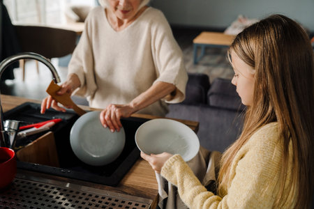 Happy grandmother and granddaughter washing dishes together in kitchen at homeの写真素材