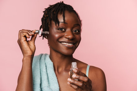 Black shirtless woman with towel smiling and applying face serum isolated over pink backgroundの写真素材
