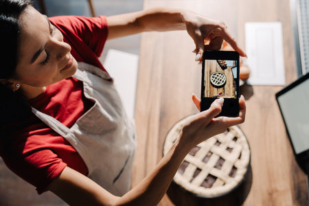 Young hispanic brunette woman wearing apron taking photo of meal while cooking in kitchen at homeの写真素材
