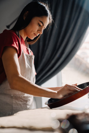 Young hispanic brunette woman wearing apron cooking and holding baking pan in kitchen at homeの写真素材