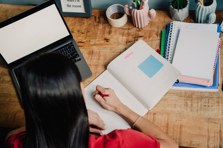 Young hispanic woman writing down notes at organizer while working with laptop at home