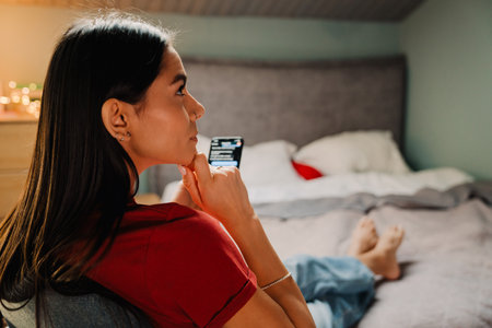 Young hispanic woman using cellphone while sitting on chair in bedroom at homeの写真素材