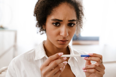 Young black woman being upset while holding pregnancy test sitting on sofa at homeの写真素材