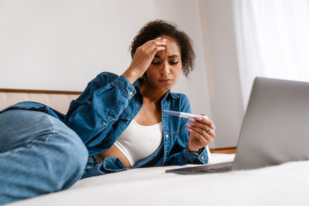 Young black woman holding pregnancy test and using laptop while lying in bed at homeの写真素材